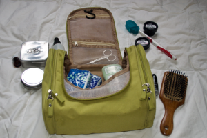 A full green toiletry bag with two tins and two bottles to the left of it, and a hair brush and hair oil bottle, plus a toothbrush, and two containers of toothpaste tabs to the right of it.