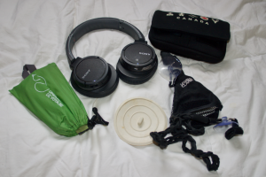 A deflated travel pillow, over ear wireless bluetooth headphones, an electronics bag stuffed full, a sink plug, and a travel clothesline; all laid out in a circle.