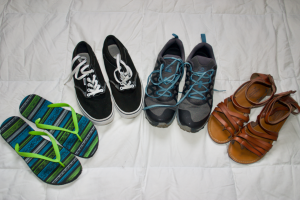 Four pairs of shoes in a semi-circle - from left to right: blue and green flip flops, black and white vans sneakers, grey hiking shoes with blue laces, and brown scrappy sandals.