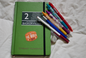 A green hard cover journal style book with Hound of the Baskervilles locations printed on the front and a "Life is short and the world is wide" sticker. Laying against the journal are a pen, a mechanical pencil, a yellow pen, a purple pen, a teal pen, and a red pen. A white eraser sits below the lowest pen.