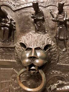 Lion head door knocker on bronze door, surrounded by engraved patterns, humans, buildings, and animals